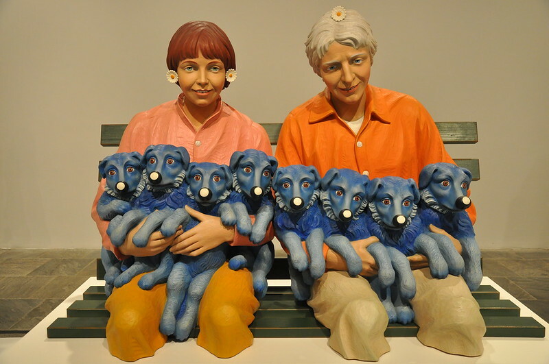 A sculpture by Jeff Koons featuring a Caucasian man with short gray hair and a Caucasian woman with short reddish hair and bangs, both wearing orange button-down shirts and small white flowers with yellow centers in their hair. They are sitting on a bench, holding eight blue puppies with white dots on their noses.