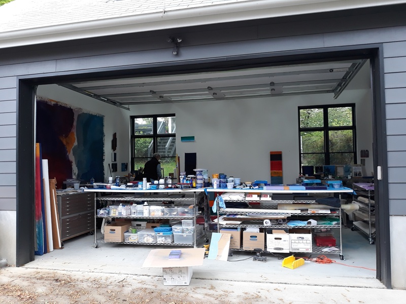 The image provides a view into the interior of McCain McMurray's art studio, taken from outside looking in through an open garage-style door. Inside the studio, there's an abundance of natural light coming through large windows. The space is organized with shelves and tables covered with numerous paint containers and art supplies, reflecting a busy and creative work environment. Large abstract paintings can be seen leaning against the walls, featuring vibrant colors and bold forms. 