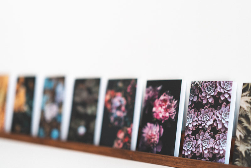 A blurred image of a row of art prints displaying various artistic designs propped on a wooden shelf, with a focus on their vivid colors and the suggestion of floral motifs