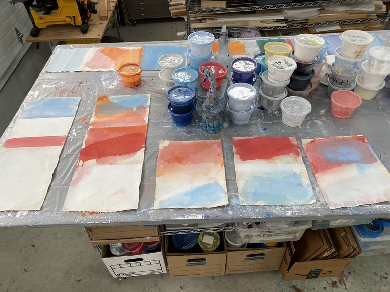 The image is taken inside McCain McMurray's studio, showcasing a series of abstract paintings in progress laid out on a worktable. The artworks are characterized by their vibrant blocks of orange, blue, and red, interspersed with softer tones and white spaces, which create a rich tapestry of color. Visible in the background are various containers of paint in a wide spectrum of colors, suggesting the artist's palette