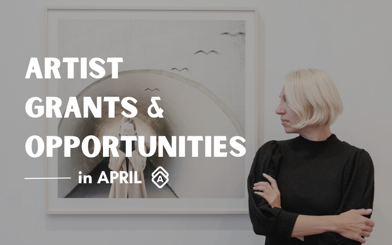 The image features bold white text overlayed that reads "ARTIST GRANTS & OPPORTUNITIES in APRIL" with the white Artwork Archive logo. In the foreground, artist Dasha Pears, with short blonde hair, dressed in a black turtleneck, stands with her arms crossed. She appears contemplative or analytical assessing the artwork. The artwork in the background, framed and mounted on the wall, shows a reflective surface with a distorted figure and what appears to be birds flying in the reflection.