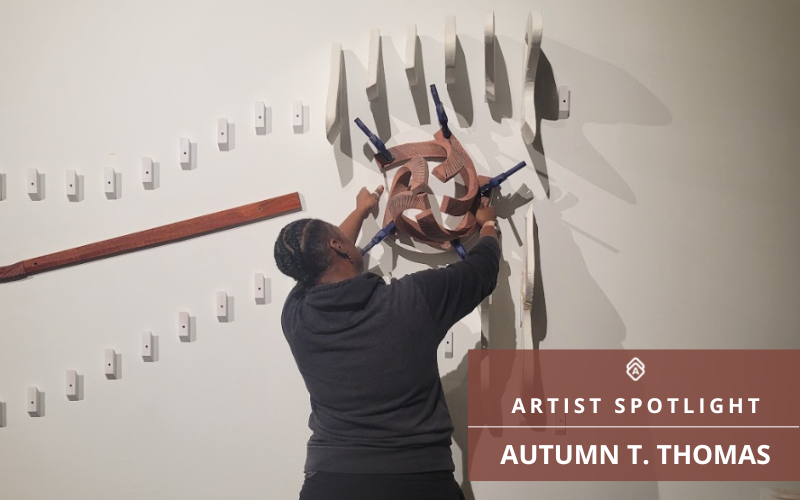 The image features Autumn T. Thomas engaged in the process of creating or assembling a wood-based art installation on a white wall. The artwork is three-dimensional, with parts of it being held together by blue clamps, suggesting it may still be under construction. Various shades and tones of the wooden pieces contribute to a dynamic and textured composition, and the shadows cast on the wall enhance the three-dimensionality of the work.  The scene is overlaid with text that reads "Artist Spotlight Autumn T. Thomas." The White Artwork Archive logo is pictured directly above the text