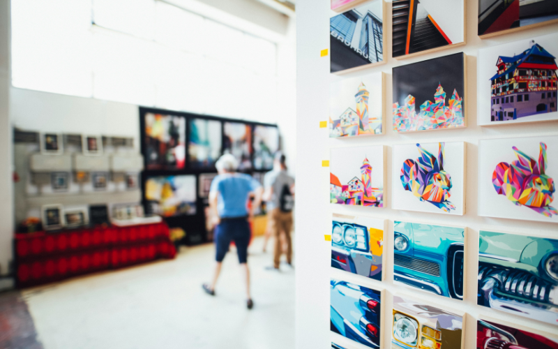 A picture from an art fair. There is a section of a wall filled with square colorful art prints. That is the only part of the image that is in focus. Beyond that, you can see a blurred view of a person in a blue shirt and dark capris observing another wall filled with art. 