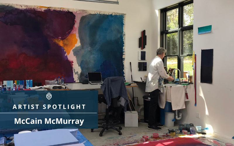 This image shows an artist's studio with a focus on a large, colorful abstract painting on the wall. Artist McCain McMurray, with his back turned to the viewer, is working at a sink, possibly cleaning brushes or mixing colors. To the left of the painting, there's a laptop on a desk, surrounded by various art supplies. Natural light streams in through a large window, casting a warm glow on the room. The walls are adorned with other smaller, abstract pieces. The text overlay reads "ARTIST SPOTLIGHT McCain McMurray,