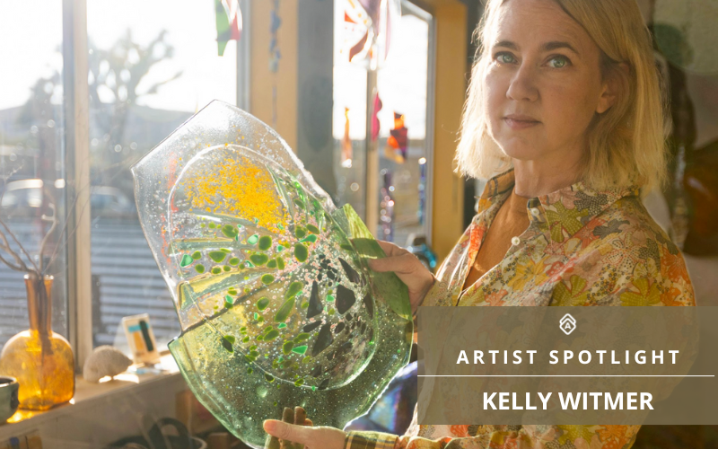 Image of the artist Kelly Witmer holding one of her glass artworks, with the caption 'Artist Spotlight - KELLY WITMER'. Kelly is pictured in her studio, partly in profile, gazing thoughtfully to the side. She holds a large, transparent glass piece with splashes of yellow and green, and an array of bubbles captured within the glass, symbolizing her intricate work with transparency and color. The background shows a sunlit room with blurred details of other art pieces and studio items. The light streaming through the window highlights the textures and colors of the glasswork