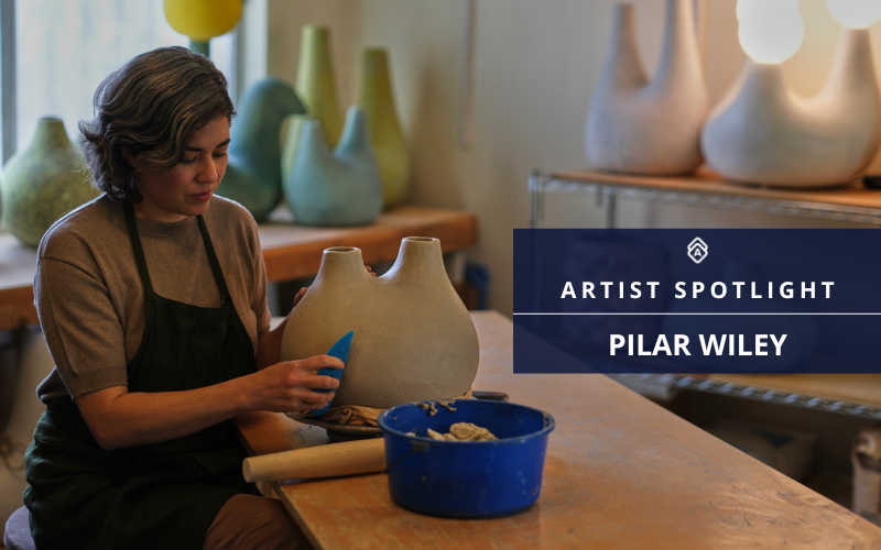 The image features artist Pilar Wiley focused on crafting a large clay pot in a studio setting. She's wearing a grey shirt and a dark apron, using a blue tool to refine the surface of the pot. In the background, shelves display various finished ceramic pieces in green and neutral tones. The image includes text that reads "Artist Spotlight: Pilar Wiley