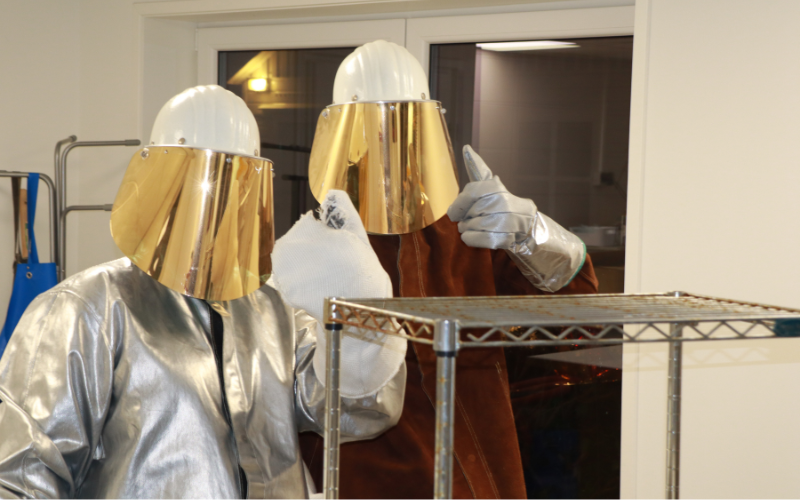 Two people wearing white protective jumpsuits and gold-colored full-face masks giving thumbs up.