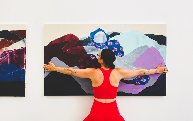 Artist Mel Reese standing in front of her painting, arms outstretched as if embracing the artwork, wearing a bright red athletic sports bra and shorts.