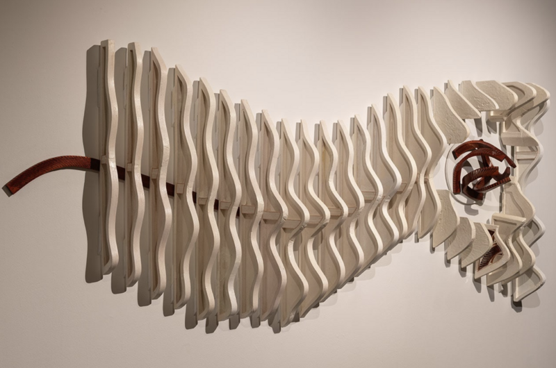 The image showcases a wood sculpture titled "In the Melody of Its Own Becoming" by Autumn T. Thomas. The sculpture features a series of undulating wooden forms that create a sense of rhythm and movement along the wall. The wooden pieces are light in color and seem to ripple outward from a central point, with some elements extending further out than others, giving the artwork a dynamic and organic feel. On the left side, a darker piece of wood with a smooth, curved surface contrasts with the lighter tones and textures. The sculpture measures 54 x 115 x 23 inches