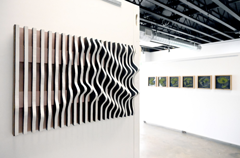 he image shows an art gallery setting with a prominent wood sculpture titled "We Would Rather Move Through Our Lives like the Flow of Water" by Autumn T. Thomas. The sculpture consists of parallel wooden slats arranged on a wall, with alternating slats protruding at varying depths to create a wavy, fluid pattern reminiscent of flowing water. The natural wood tones contrast with the white wall. To the right of the sculpture, a series of framed artworks are displayed in a row. The sculpture measures 23 x 35 x 4 inches