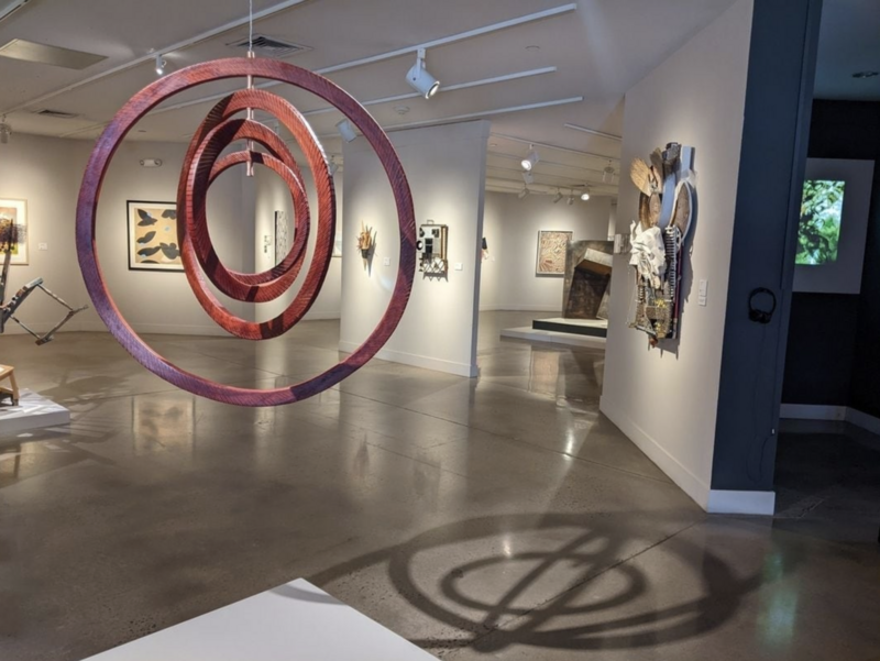 The image shows a contemporary art gallery with various artworks on display. The focus is on a striking hanging sculpture titled "Eccentricity" by Autumn T. Thomas, which features concentric wooden rings suspended in the air. The rings have a reddish finish that gives them a rich, warm hue. The piece is dimensionally listed as 36 x 36 x 36 inches. The sculpture casts an interesting shadow on the polished gallery floor. Around the sculpture, the walls are adorned with a diverse array of art pieces in different styles and mediums.