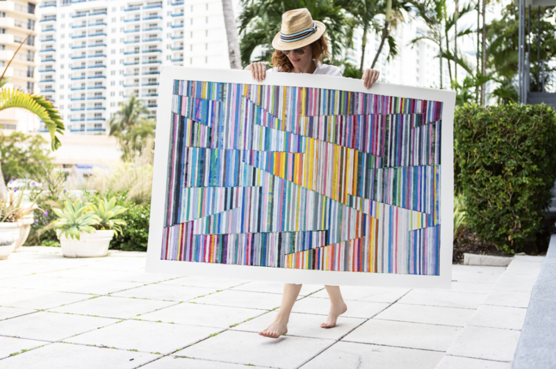 The image features a woman, identified as the artist Johanna Boccardo, holding a large, colorful abstract painting. She is outdoors, in a garden area with high-rise buildings in the background, indicative of an urban setting. The artist is partially obscured by the artwork, with only her face, arms, and lower legs visible. She is wearing a straw hat and is barefoot on the paving stones. The painting has a vibrant mix of multicolored vertical stripes, interrupted by angled, geometric breaks in the pattern. The setting is bright and sunny