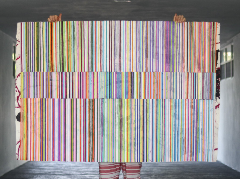 The image shows a colorful abstract painting held up by an artist. The artwork is dominated by vertical stripes in various colors creating a vibrant, rainbow-like effect. The stripes are interrupted by three horizontal bands where the colors are disconnected, creating a sort of optical illusion. The hands of the person holding up the painting are visible at the top, and the lower part of the person's body can be seen at the bottom, wearing striped pants that mimic the painting's pattern. The artwork is titled "Beach Blanket 1" and measures 43 x 63 inches, created by Johanna Boccardo.