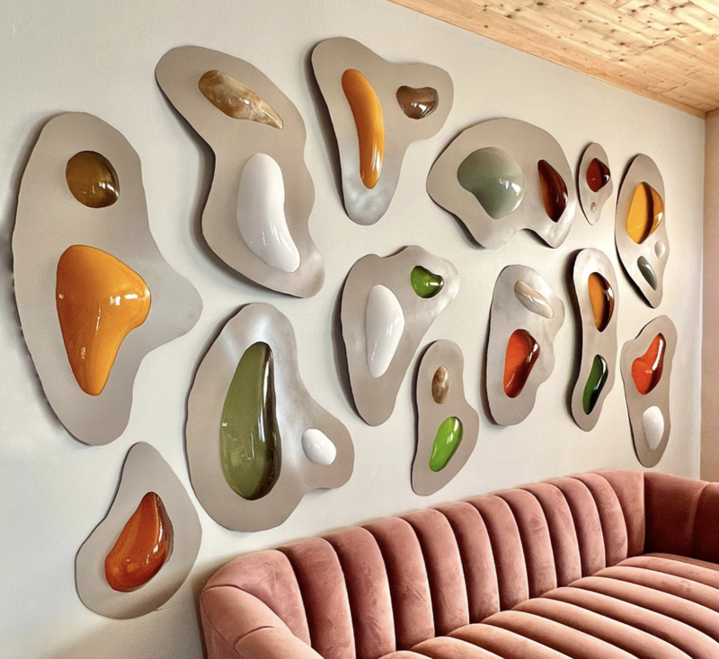 A photograph of a modern art installation titled 'Custom Series—Kuwait Project' by Kelly Witmer. The artwork features an arrangement of variously shaped and sized abstract forms on a wall, each form resembling a fluid or organic shape with smooth, rounded edges. These shapes are rendered in a glossy finish, showcasing a palette of earthy and vibrant colors such as green, orange, brown, and white. The forms are set against a neutral wall and are interspersed in a seemingly random, yet harmonious composition. Below the installation is a plush, pink, tufted couch. The piece is listed as measuring 70 x 120 x 10 inches and was created in 2023.