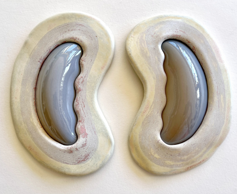 Image of a pair of ceramic artworks by Kelly Witmer, titled 'Gray and Cream Pair'. The two pieces are almost identical, featuring smooth, bean-like shapes in gray and cream hues. Each form has a glossy gray center that is kidney-shaped, surrounded by a thick, cream-colored border with subtle hints of red and yellow. The textures and colors suggest depth and dimension, with the pieces casting soft shadows on the white wall behind them. This artwork measures 9 x 20 x 2 inches and was created in 2024.