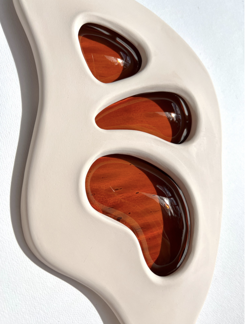 A close-up photo of a sculptural artwork titled 'Amber Tears' by Kelly Witmer. The piece features three glossy, amber-colored, teardrop-shaped insets nestled within a curvaceous, matte-finished, off-white ceramic frame. The rich, translucent amber shapes cast warm reflections and give the impression of depth against the solid, white background. The piece measures 11 x 8 x 3 in, and was created in 2022