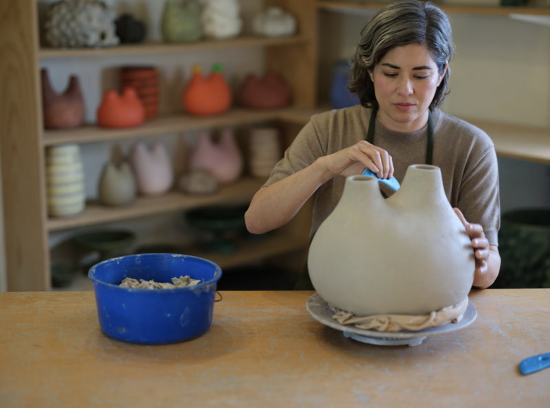  The image shows a woman in a pottery studio, meticulously working on a large, clay pot. She is using a small blue tool to shape or decorate the pot. She wears a grey top and a dark apron. In the foreground, there's a blue bowl containing clay scraps on a table. In the background, shelves filled with various colorful pottery pieces are visible. The caption notes the artist as Pilar Wiley, with photo credit to Reynaldo Rivera, and mentions the photo is courtesy of the artist.