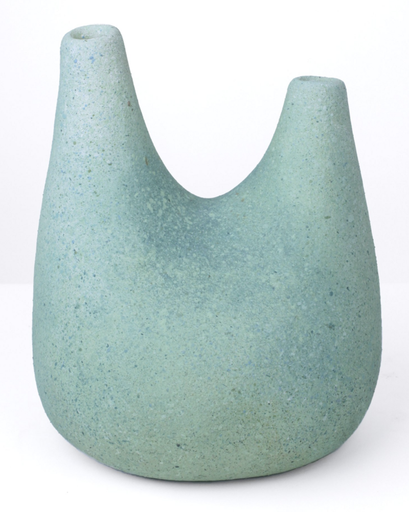 The image shows a close-up of a ceramic vase with a unique, curved neckline design. The vase has a smooth surface and a pale green color with subtle speckles distributed throughout. The shape is asymmetrical, with one side rising taller than the other, creating a distinctive silhouette. The piece was created by Pilar Wiley, it titled 'Seafoam', and measures 13.5 x 11.25 x 10.25 in, made in 2023