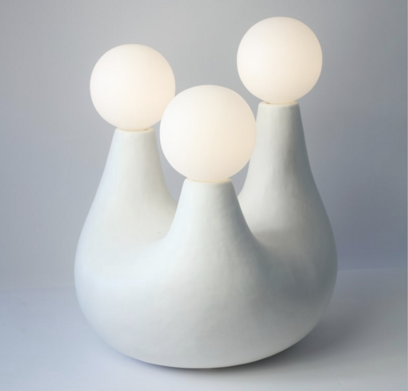  The image displays an artistic light installation titled "Light Triad" by Pilar Wiley, created in 2023. It features three pale, curved ceramic bases, each supporting a spherical, illuminated globe on top. The artwork measures 22.625 x 18.5 x 18 inches. The ceramics have a matte finish in a soft, light gray tone, contributing to a minimalist and modern aesthetic. 