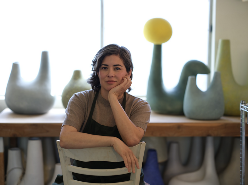 The image features artist Pilar Wiley seated at a table in a ceramic studio. She is leaning on her left arm, resting her chin on her hand, and looking directly at the camera with a gentle expression. She wears a grey shirt and a dark apron. In the background, several large ceramic pieces are displayed on a shelf by a window, including a distinctive piece with a yellow spherical top. The studio appears bright and airy, emphasizing a creative and artistic environment.